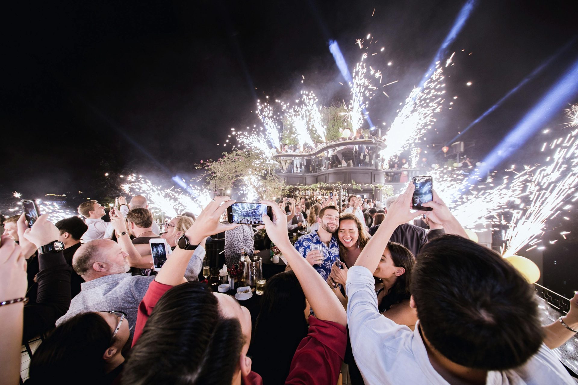 Vibrant party scene with fireworks and joyful crowd capturing moments on their phones.